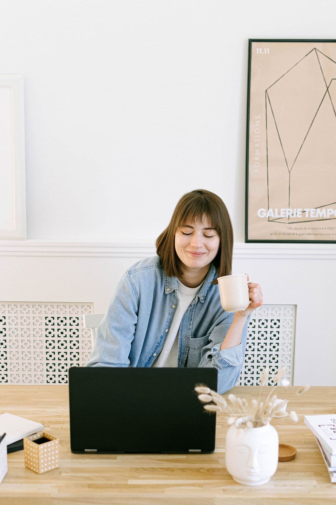 A woman smiling over a cup of coffee at her desk, with a laptop in front of her